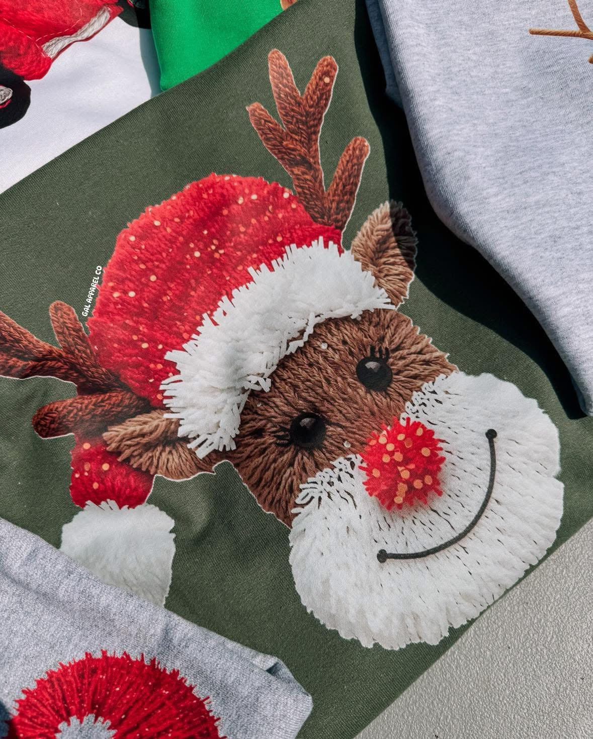 Reindeer faux yarn design on Christmas t-shirt and sweatshirt, featuring red hat and pom pom nose festive holiday graphic.
